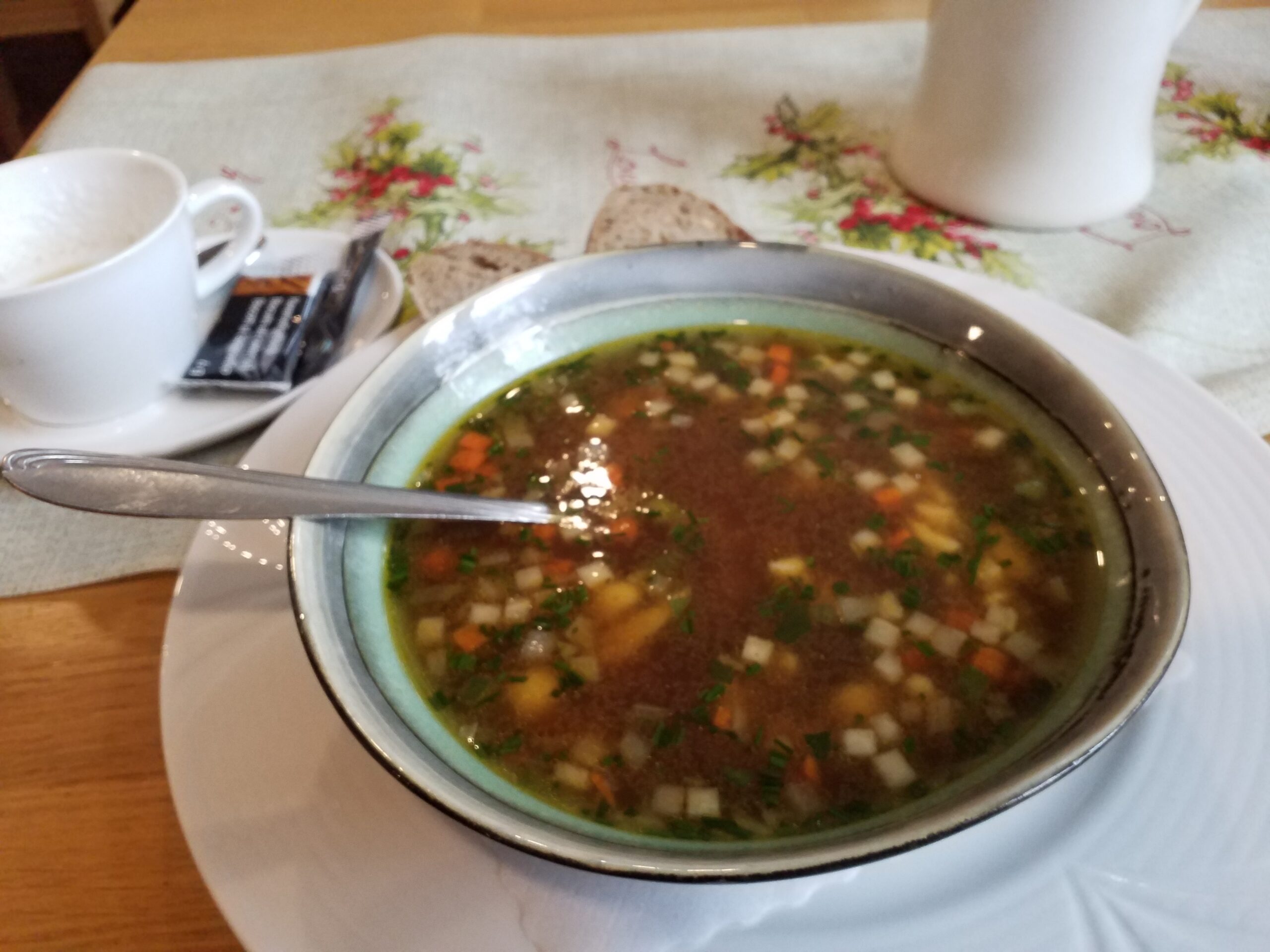Teller warme klare Suppe mit Gemüsewürfelchen, Scheibe Brot daneben, Tischdecke, links Kaffeetasse.