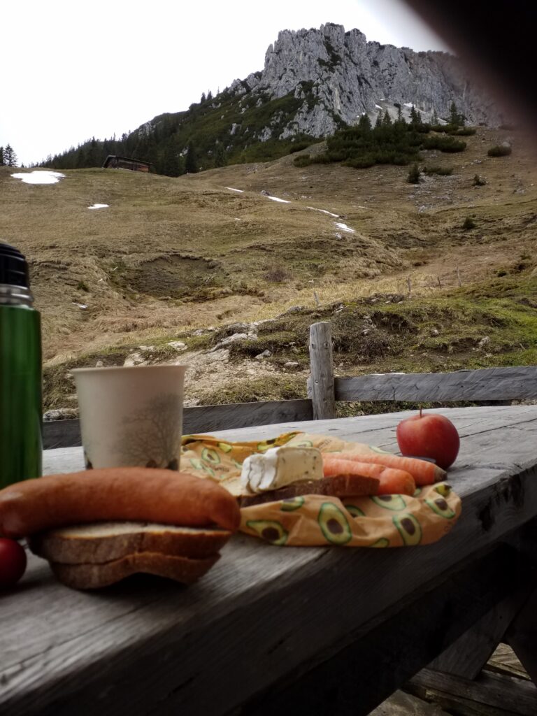 Vorne ein Holztisch mit Apfel, Brot und Käse auf einem Tuch, Trinkbecher, Trinkflasche. Dahinter führt der Weg zu einem kaum sichtbaren Häuschen. Im Hintergrund das felsige Bergmassiv.