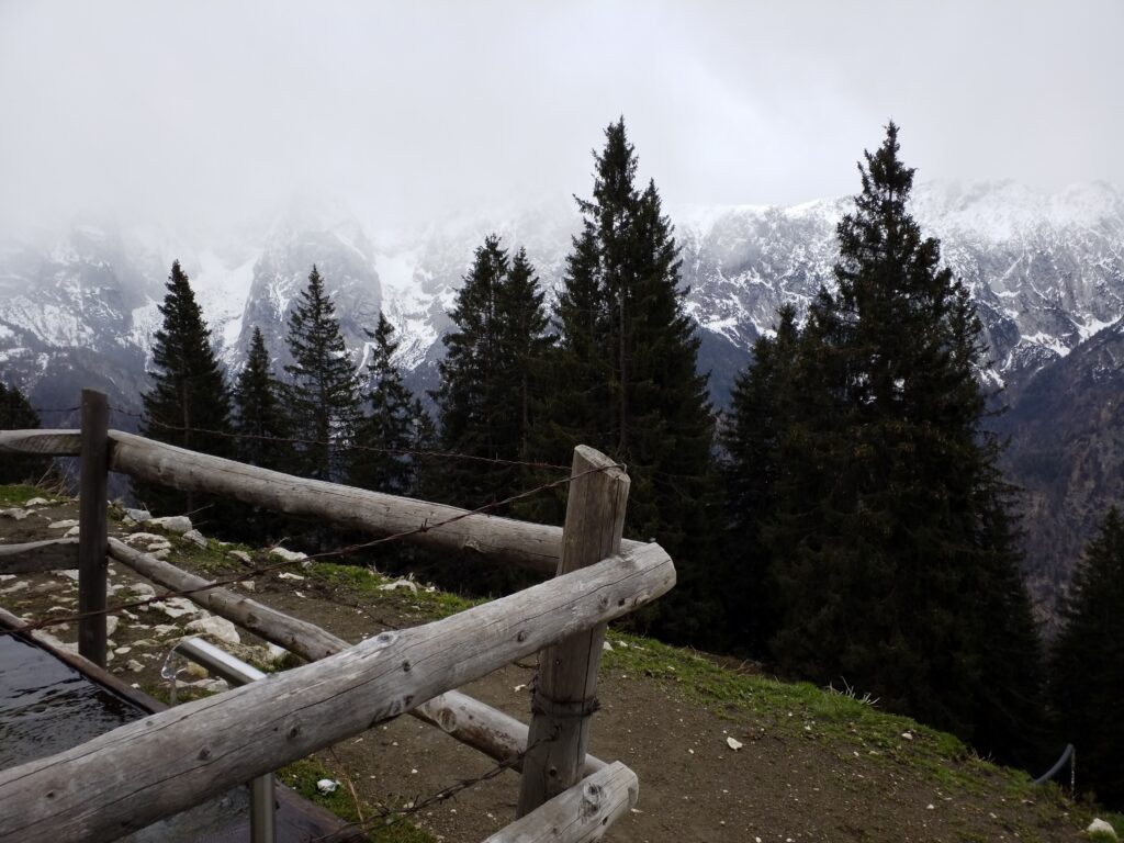 Links Holzzaun mit Stacheldraht oben. Er umgibt eine Wasserstele. Der Weg führt an einem steilen Wiesenhang um die Wasserstelle. Nadelbäume. Dahinter ist das Gebirge zu sehen. Leicht schneebedeckt.