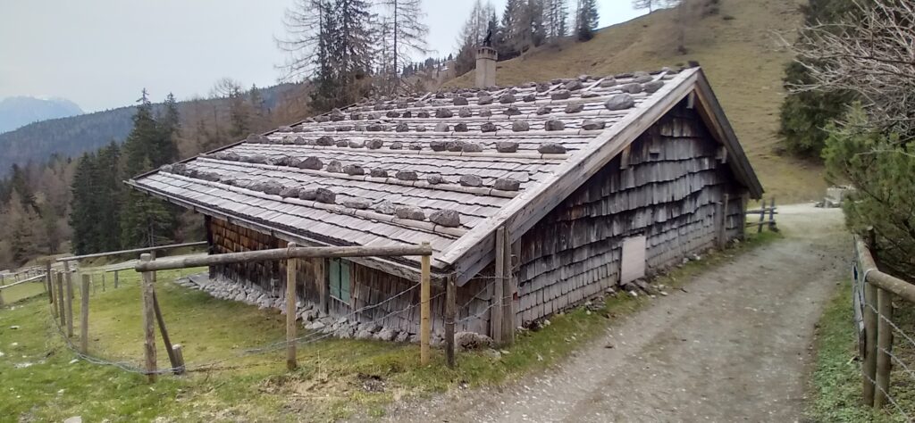 niedriges Holzschindelhaus im Gebirge auf einer Wiese, holzumzäunt. Das Dach ist mit Reihen von Steinen gesichert.