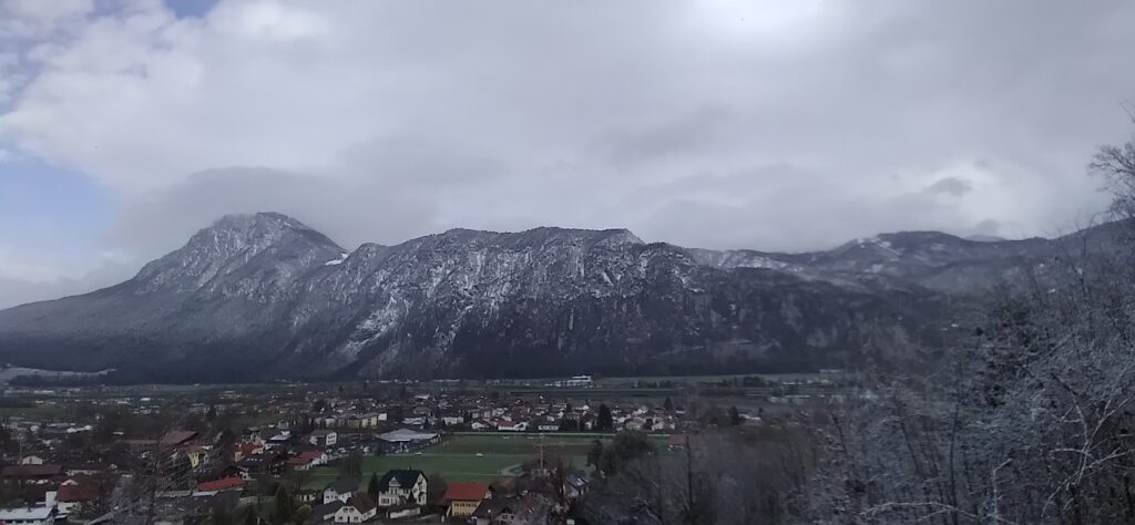 Weites Tal mit der Stadt Kiefersfelden, dahinter das große Massiv des Kaiser von rechts schneebepuderte Zweige. bedeckter Himmel.